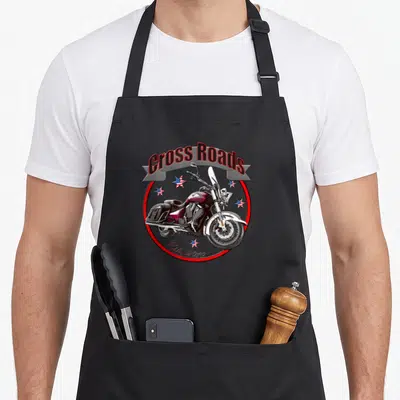 Tablier de cuisine noir avec illustration d'une moto cruiser bordeaux, étoiles US et texte Cross Roads Est. 2010 dans un cercle rouge.