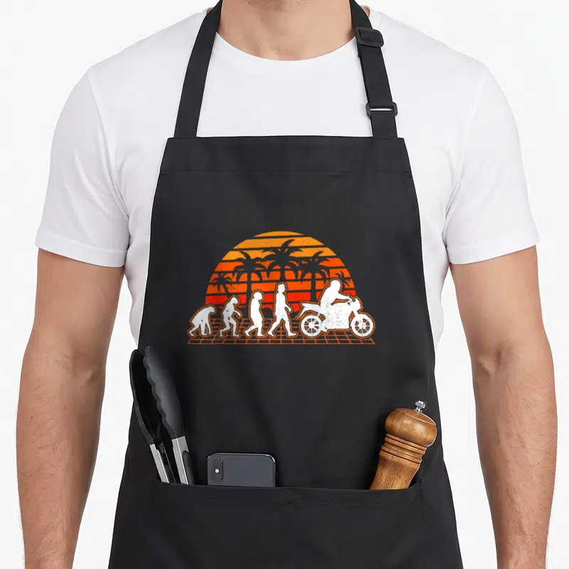 Tablier de cuisine noir avec illustration évolution humaine terminant par un motard sur fond de soleil orange et palmiers rétro.