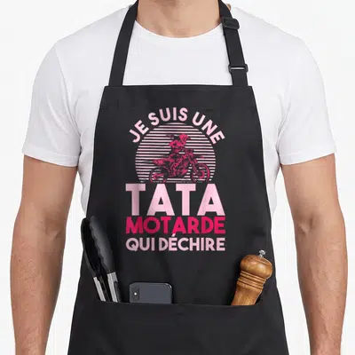 Tablier de cuisine noir avec inscription Je suis une tata motarde qui déchire et silhouette de moto rose sur fond strié.