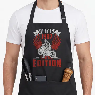 Tablier de cuisine noir avec illustration vintage d'un motard aux ailes rouges et l'année 1987 imprimée en rouge.