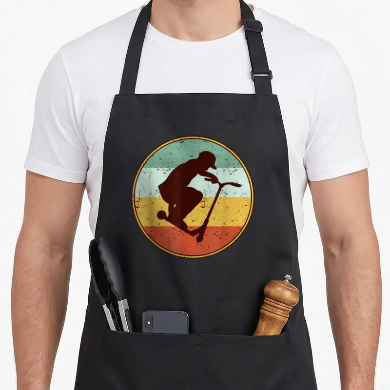 Tablier de cuisine noir avec une illustration circulaire d'un skieur de trottinette en silhouette sombre sur fond de bandes colorées délavées.