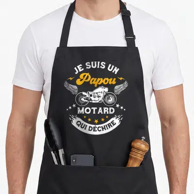 Tablier de cuisine noir avec inscription Je suis un Papou motard qui déchire et illustration de moto ailée blanche et jaune.