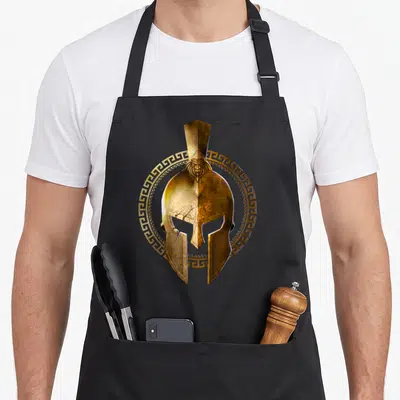 Tablier de cuisine noir avec un casque spartan doré entouré d'un cercle à motifs géométriques antiques.