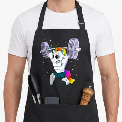 Tablier de cuisine noir avec un Manly Unicorn musclé soulevant une barre de poids colorée.