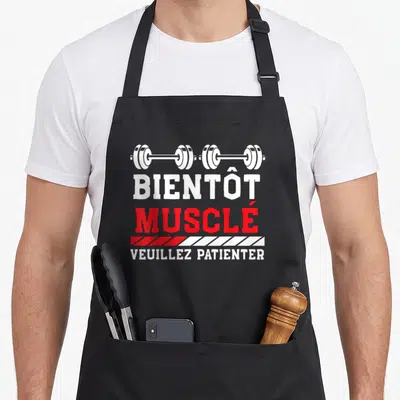 Tablier de cuisine noir avec illustration de barre de poids et texte Bientôt Musclé Veuillez Patienter en rouge et blanc.