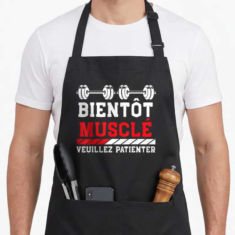 Tablier de cuisine noir avec illustration de barre de poids et texte Bientôt Musclé Veuillez Patienter en rouge et blanc.