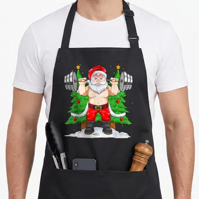 Tablier de cuisine noir avec illustration d'un Père Noël musclé soulevant une barre de poids devant des sapins.