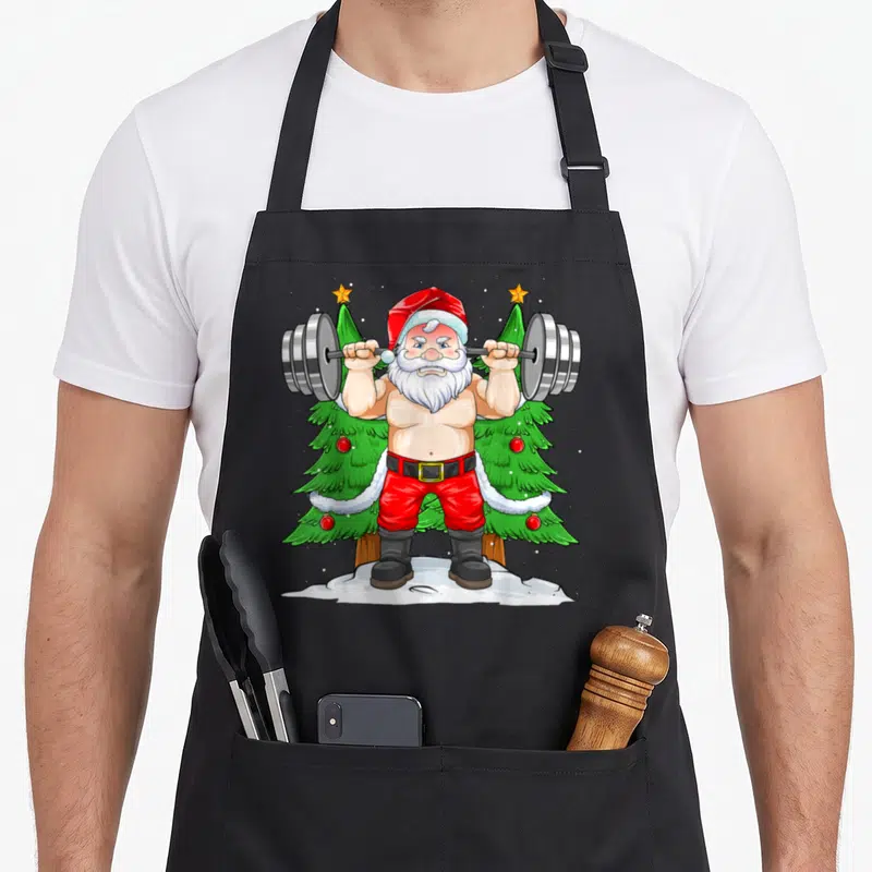Tablier de cuisine noir avec illustration d'un Père Noël musclé soulevant une barre de poids devant des sapins.