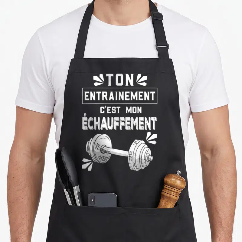 Tablier de cuisine noir avec le texte Ton entraînement c'est mon échauffement au-dessus d'une barre d'haltère blanche stylisée.