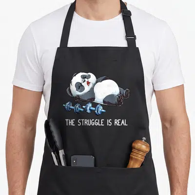 Tablier de cuisine noir avec un panda épuisé allongé à côté de deux barres bleues et le texte The Struggle Is Real en blanc.
