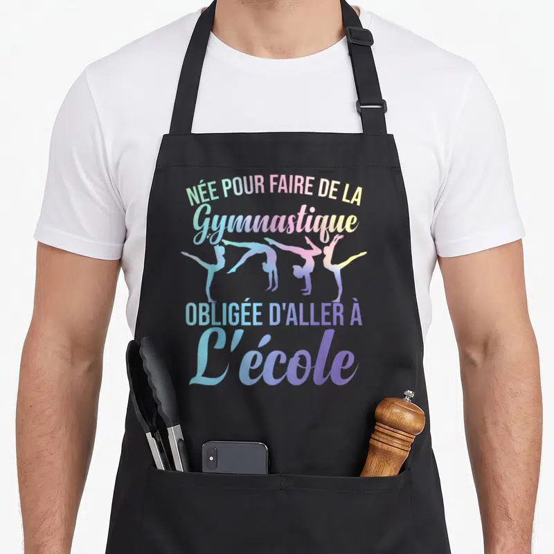 Tablier de cuisine noir avec des silhouettes de gymnastes et le texte née pour faire de la gymnastique obligée d'aller à l'école.