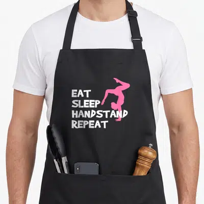 Tablier de cuisine noir avec texte blanc Eat Sleep Handstand Repeat et silhouette rose d'une personne en appui main.
