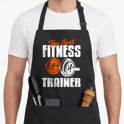 Tablier de cuisine noir avec The Best Fitness Trainer et une barre de poids orange et blanche imprimée au centre.