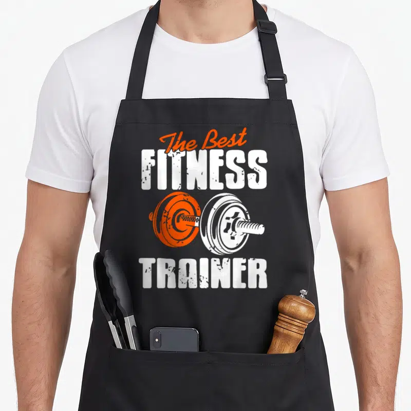 Tablier de cuisine noir avec The Best Fitness Trainer et une barre de poids orange et blanche imprimée au centre.