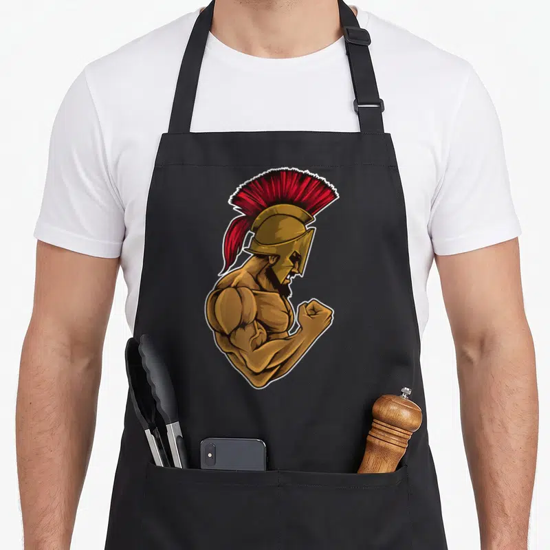 Tablier de cuisine noir avec illustration d'un guerrier spartain musclé portant un casque doré à crête rouge vif.