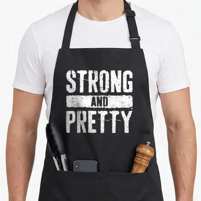 Tablier de cuisine noir avec l'inscription blanche Strong And Pretty en lettres capitales texturées.