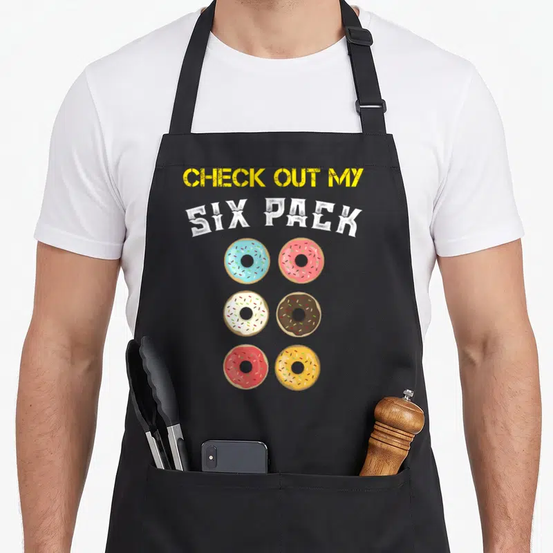 Tablier de cuisine noir avec inscription Check Out My Six Pack et six donuts colorés disposés en grille.
