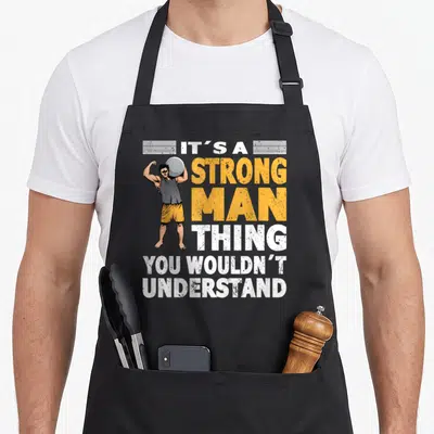 Tablier de cuisine noir avec illustration d'un homme fort tenant une boule de et le texte It's a strong man thing you wouldn't understand.