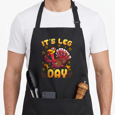 Tablier de cuisine noir avec un dindon cartoon courant et le texte It's Leg Day entouré de feuilles d'automne.