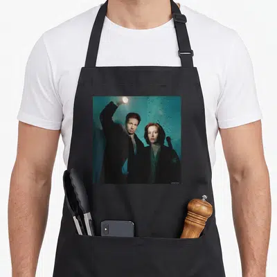 Tablier de cuisine noir avec illustration de David Duchovny et Gillian Anderson sous une lumière de lampe torche.