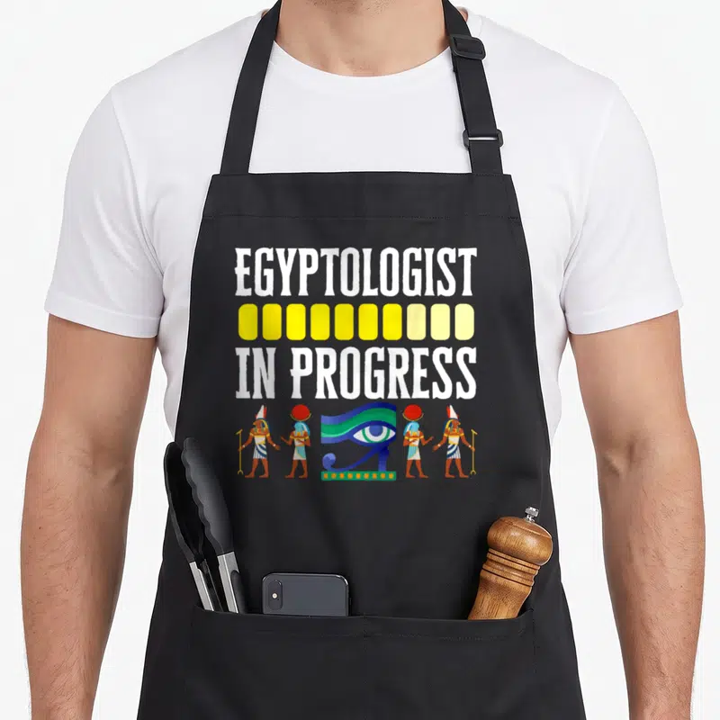 Tablier de cuisine noir avec inscription Égyptologiste in progress et symboles pharaoniques colorés.