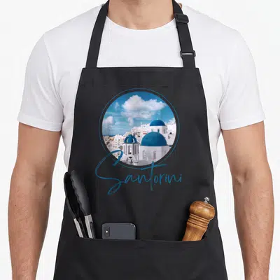 Tablier de cuisine noir avec un motif circulaire représentant les dômes bleus et les bâtiments blancs de Santorini sous un ciel nuageux.
