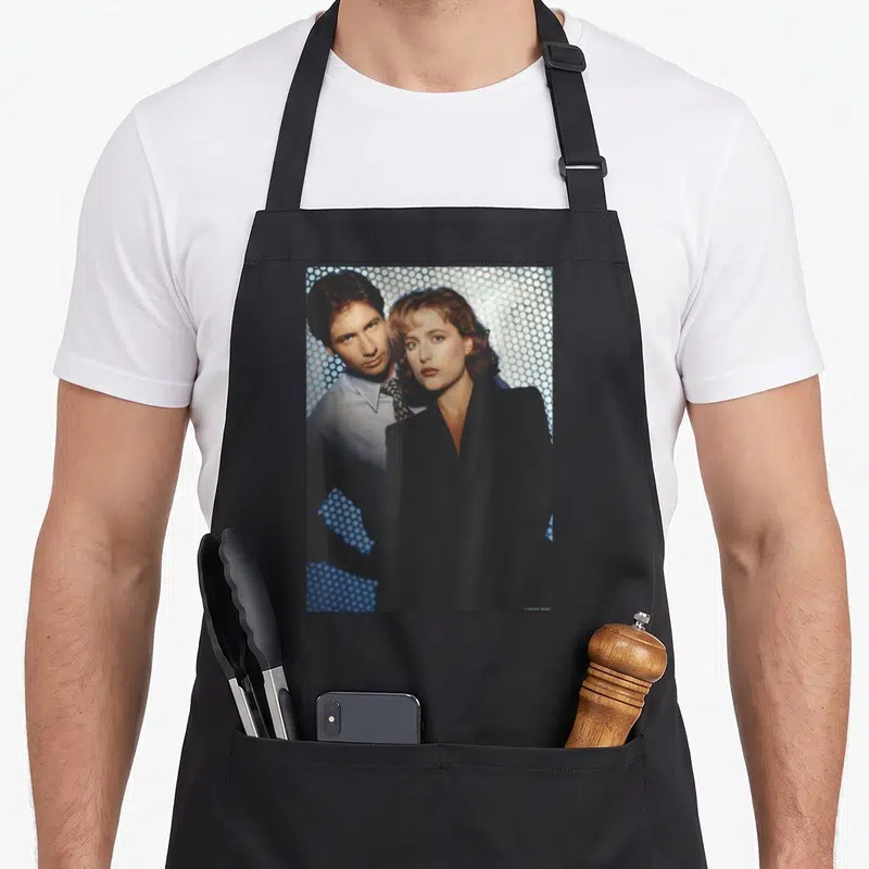 Tablier de cuisine noir avec David Duchovny et Gillian Anderson devant un motif de points bleus.