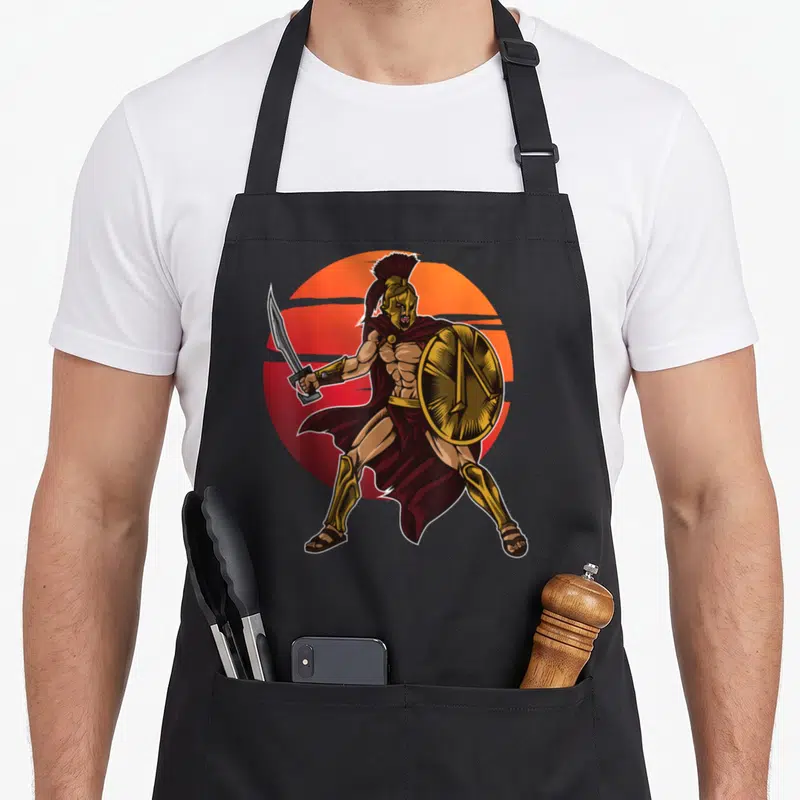Tablier de cuisine noir avec illustration d'un guerrier spartiate au casque doré et bouclier circulaire.