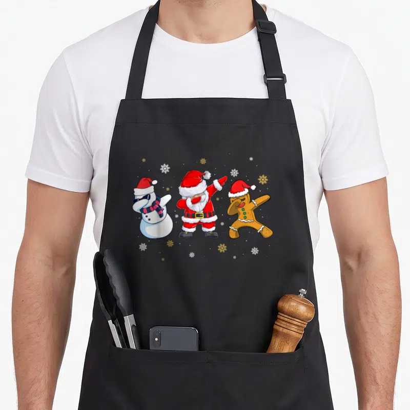 Tablier de cuisine noir avec Père Noël, bonhomme de neige et biscuit de Noël en train de faire le dab au milieu de flocons de neige.