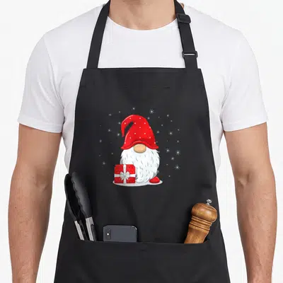 Tablier de cuisine noir avec illustration d'un gnome au chapeau rouge tenant un cadeau sous une inscription festive.