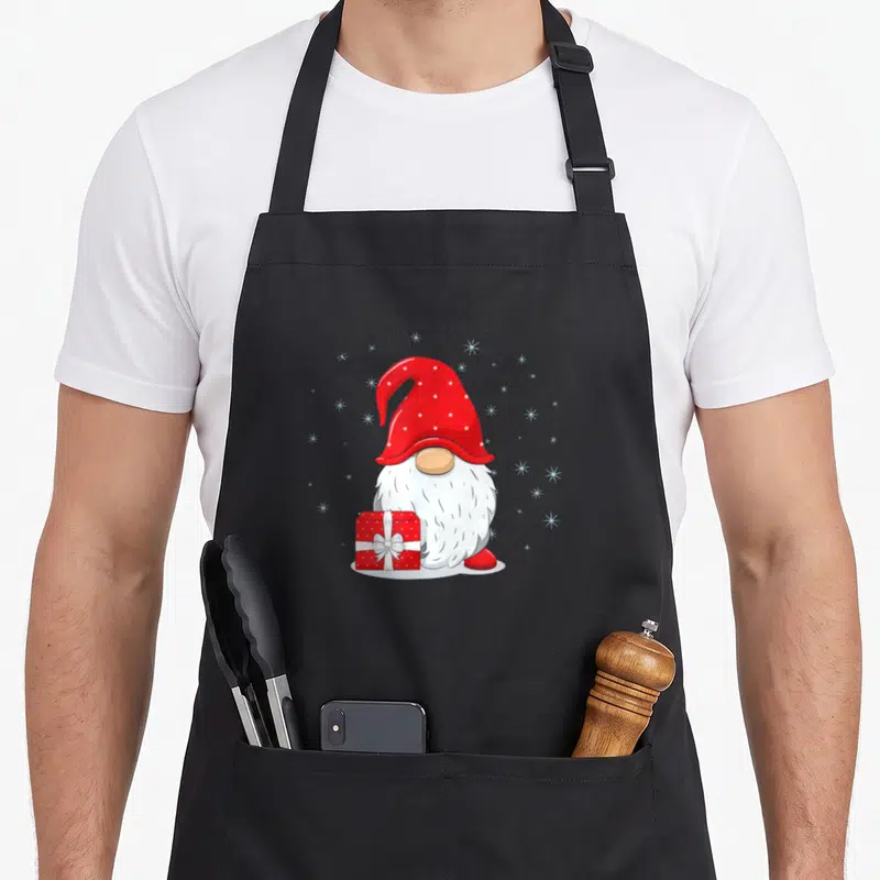 Tablier de cuisine noir avec illustration d'un gnome au chapeau rouge tenant un cadeau sous une inscription festive.