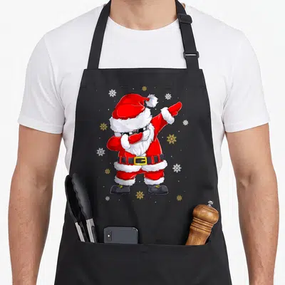 Tablier de cuisine noir avec illustration d'un Père Noël effectuant une pose dab sous des flocons de neige dorés et blancs.
