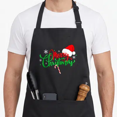 Tablier de cuisine noir avec inscription Merry Christmas, bonnet rouge, flocons blancs et canne à sucre bicolore.