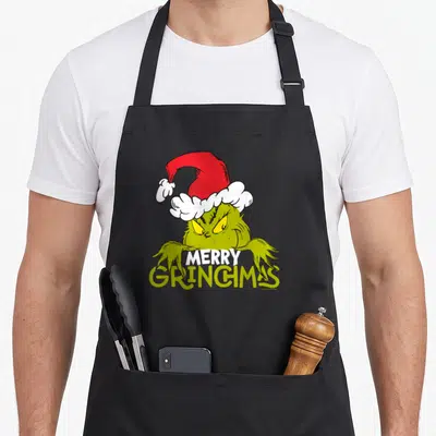 Tablier de cuisine noir avec le Grinch en bonnet rouge et l'inscription Merry Grinchmas imprimée au centre.