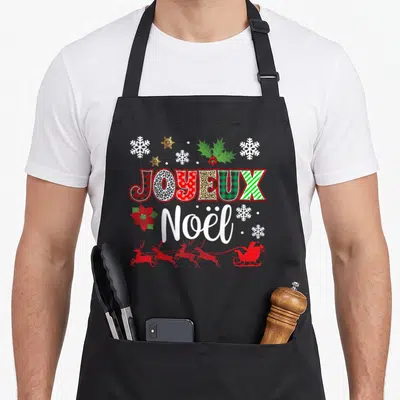 Tablier de cuisine noir avec l'inscription Joyeux Noël entouré de flocons, de rennes rouges et de feuilles de houx.