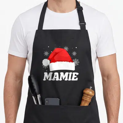 Tablier de cuisine noir avec chapeau Père Noël rouge et le mot Mamie en blanc entouré de flocons de neige stylisés.