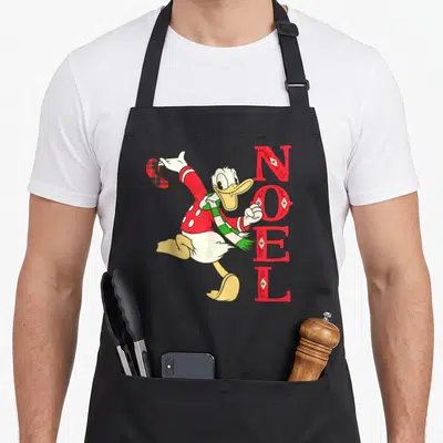 Tablier de cuisine noir avec Donald Duck en pull rouge et scarpe verte à côté du texte NOEL en lettres rouges décorées.