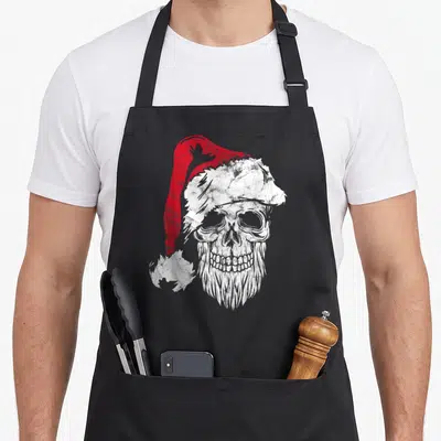 Tablier de cuisine noir avec illustration d'un crâne blanc portant un chapeau de Père Noël rouge à bordure blanche usée.