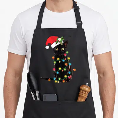 Tablier de cuisine noir avec un chat noir portant une casquette rouge et enroulé dans des guirlandes lumineuses multicolores.