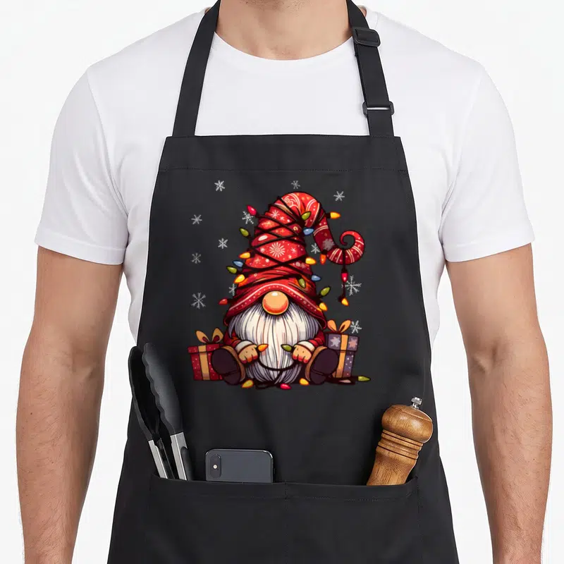 Tablier de cuisine noir avec un gnome de Noël assis entre deux cadeaux sous une guirlande lumineuse et des flocons.