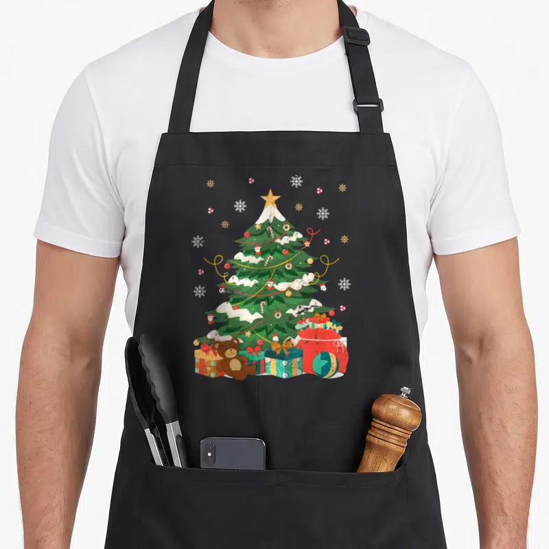 Tablier de cuisine noir avec un sapin de Noël enneigé, des cadeaux et un ours en peluche imprimés.