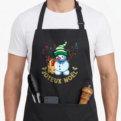 Tablier de cuisine noir avec bonhomme de neige au chapeau vert et panier de cadeaux sous l'inscription Joyeux Noël.