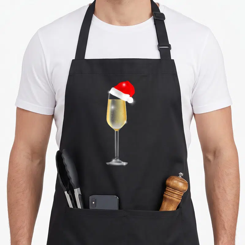 Tablier de cuisine noir avec un verre de champagne doré surmonté d'un petit bonnet rouge de Noël brillant.