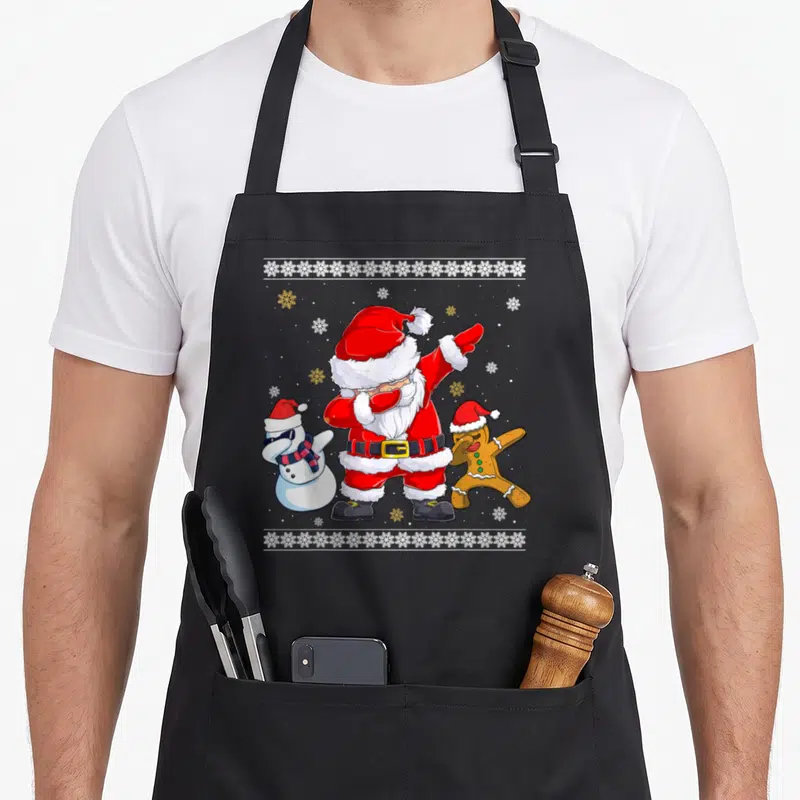 Tablier de cuisine noir avec un Père Noël faisant le dab entouré d'un bonhomme de neige et d'un biscuit de Noël.