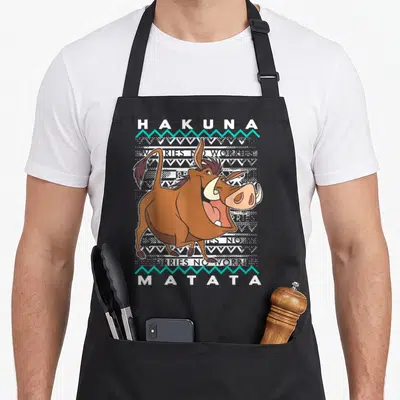 Tablier de cuisine noir avec Pumbaa souriant, inscription Hakuna Matata et motifs géométriques festifs en blanc et turquoise.