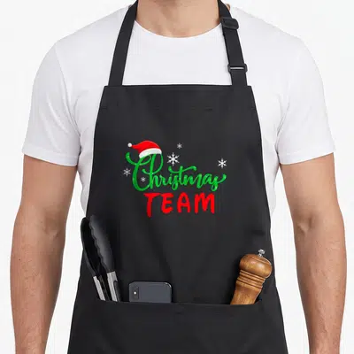 Tablier de cuisine noir avec inscription Christmas Team et bonnet de Père Noël rouge sur fond gris uni.