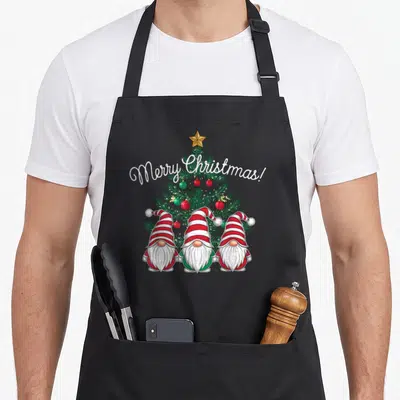 Tablier de cuisine noir avec trois gnomes à chapeaux rayés rouges et blancs devant un sapin de Noël orné de boules et d'une étoile dorée.