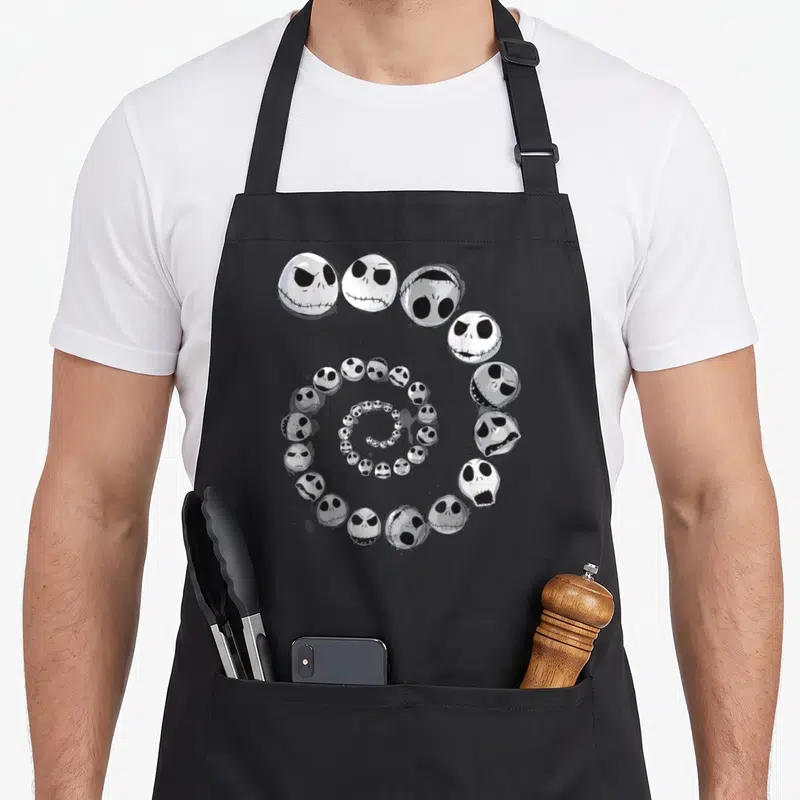 Tablier de cuisine noir avec une spirale de têtes de Jack Skellington aux expressions variées en noir et blanc.