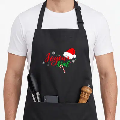 Tablier de cuisine noir avec inscription Joyeux Noël en lettres rouges et vertes, orné d'un bonnet de Père Noël et d'une canne à sucre.