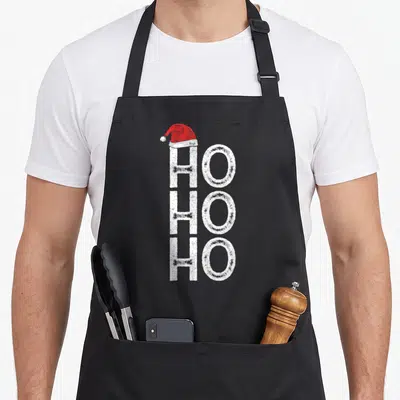 Tablier de cuisine noir avec le texte Ho Ho Ho en blanc vertical surmonté d'un bonnet de Père Noël rouge.